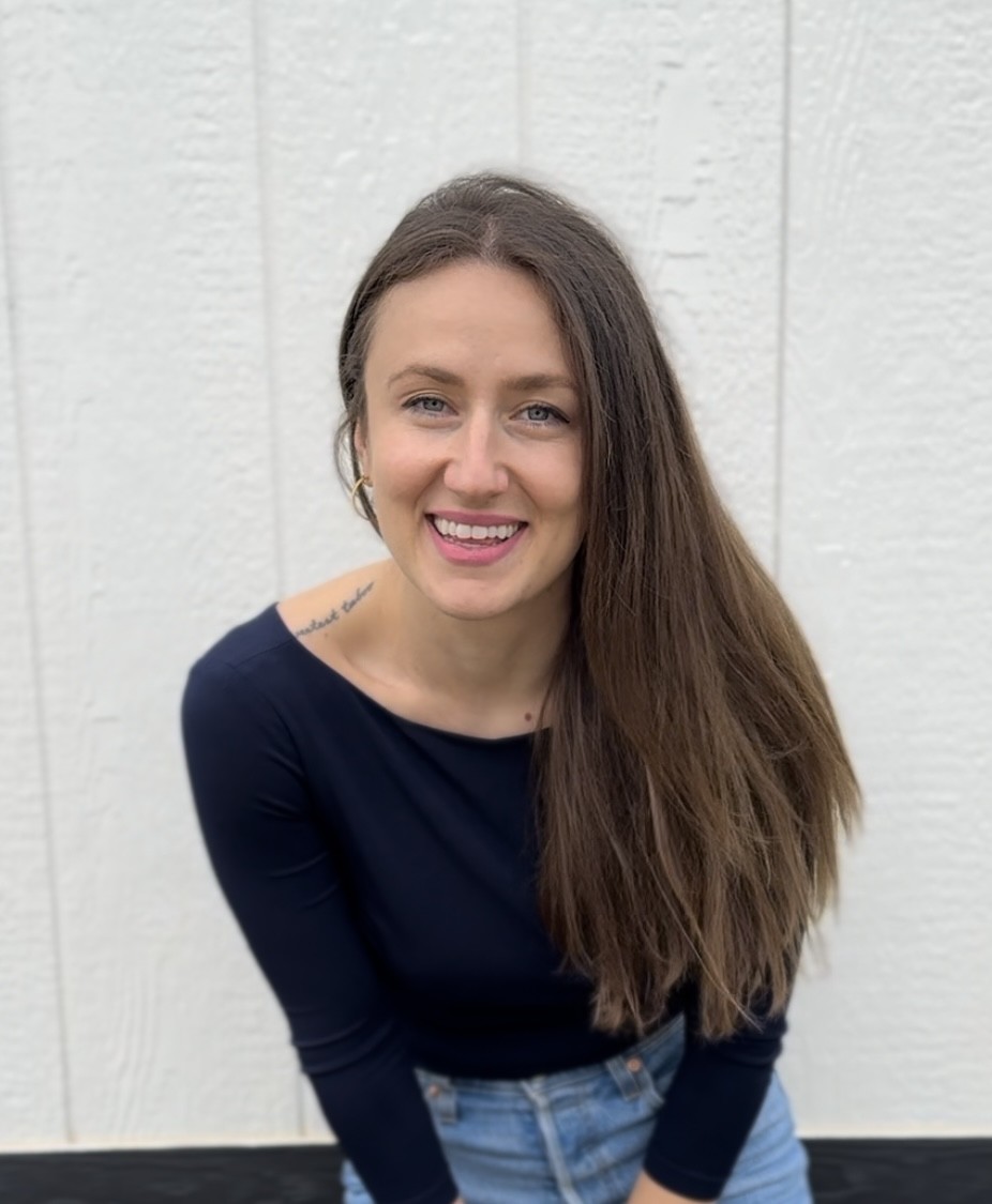 Anya Shakh smiling, with long brown hair, in a navy top against a white wall